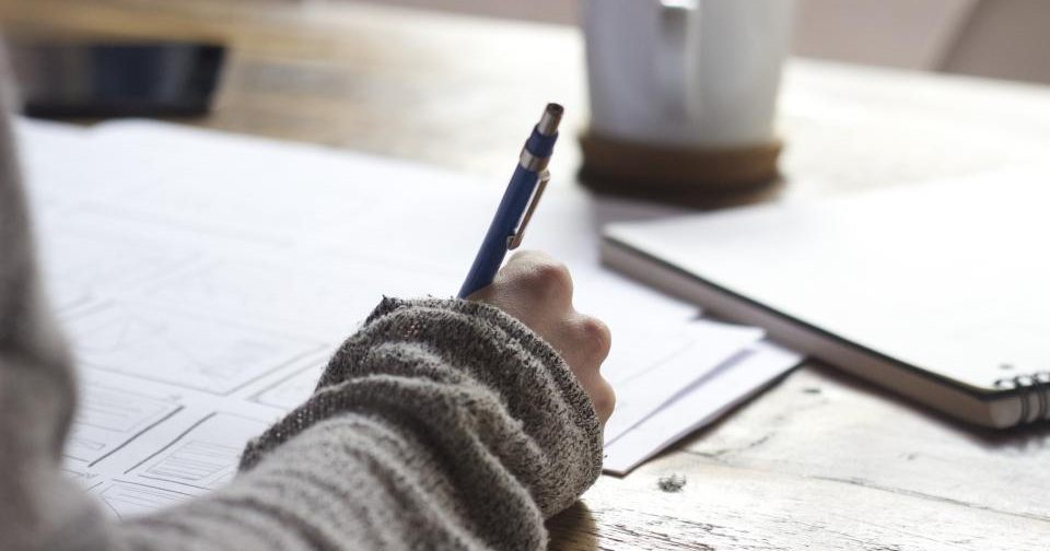 Mão a escrever notas num caderno em papel, com uma chávena de café ao fundo